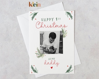 May include: A white Christmas card with a green and red floral design. The card features a photo of a father holding a baby. The text on the card reads "Happy 1st Christmas as my daddy".