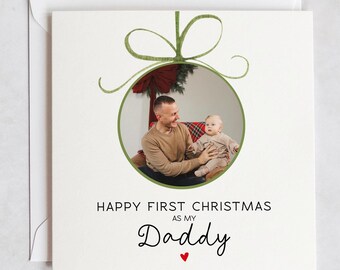 Può includere: Un biglietto di Natale con una foto di un padre e il suo bambino all'interno di un ornamento verde. Il testo recita "Buon primo Natale come mio papà".