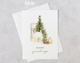 Puede incluir: Una tarjeta blanca con una ilustración de acuarela de un pequeño pueblo con un árbol de Navidad y el texto "seasons greetings".