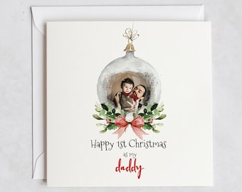 Può includere: Un biglietto di Natale con un'illustrazione ad acquerello di un ornamento argentato con una foto di un padre e un figlio al suo interno. L'ornamento è decorato con foglie verdi, bacche rosse e un fiocco rosa. Il testo "Buon 1° Natale come mio papà" è scritto sotto l'ornamento. La carta è di klein.
