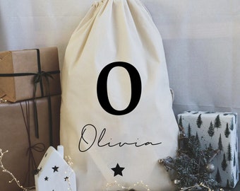 May include: A white canvas drawstring bag with a black number "0" and the name "Olivia" written in cursive. A black star is below the name. The bag is sitting on a table with other wrapped gifts.