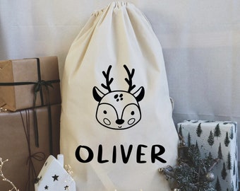 May include: A white canvas drawstring bag with a black reindeer illustration and the name "OLIVER" printed on the front. The bag is surrounded by Christmas decorations.