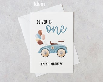 May include: A blue and white toy car with balloons attached to the back. The card says "Oliver is one" and "Happy Birthday".
