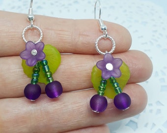 May include: Purple and green beaded earrings with a flower charm and two round beads. The earrings are silver and have a hook closure.