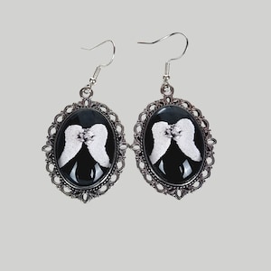 Puede incluir: Un par de pendientes plateados con un fondo negro ovalado con alas de ángel blancas. Los pendientes tienen un marco de filigrana decorativo y un cierre de gancho simple. Un accesorio de temática gótica o celestial.