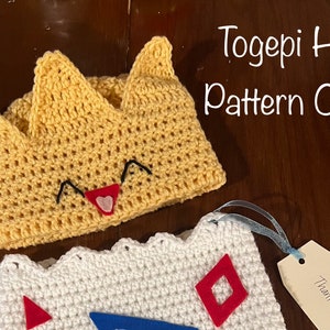May include: A crocheted Togepi hat pattern. The hat is yellow with a crown shape and a smiling face with a red heart. The text "Togepi Hat Pattern Only" is written in white on a brown background.