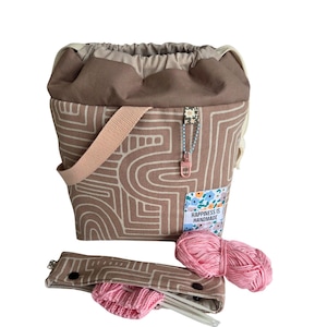 Könnte beinhalten: Eine braune und cremefarbene Garn-Projekt-Tasche mit geometrischem Muster, einem rosa Riemen und einem Kordelzugverschluss. Die Tasche hat ein kleines Etikett mit der Aufschrift "Happiness is Handmade". Ein passendes Etui enthält Stricknadeln und einen kleinen rosa Strickartikel, mit zwei Knäueln rosa Garn.