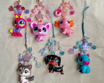 May include: A collection of colorful animal-shaped charms with beaded accents and attached lanyards. The charms include a pink koala, a pink axolotl, a black and brown dachshund, a blue hippo, a husky, and a multi-colored bug.