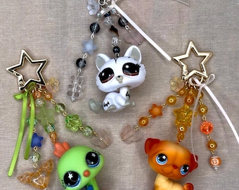 May include: Three colorful keychains featuring animal figurines. The keychains have star-shaped and round gold-tone clasps, with beaded accents in shades of green, orange, and clear. One keychain has a white cat figurine, another a green creature, and the last an orange dog.