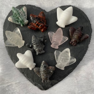 May include: A collection of ten bee-shaped crystal carvings in various colours, including green, brown, white, pink, and grey. The carvings are arranged on a heart-shaped black slate surface.