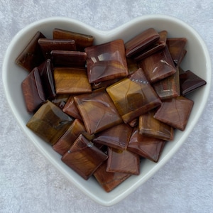 May include: A bowl filled with polished tiger's eye stones, each stone is a square shape with a golden brown colour and a reddish brown hue.