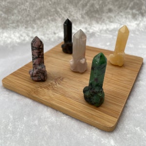 May include: Six polished crystal points in various colors, including pink, black, white, green, and yellow, are arranged on a wooden surface.