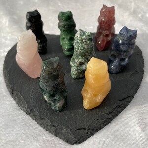 May include: A selection of small owl figurines carved from gemstones like rose quartz, obsidian, and jade. The owls are displayed on a heart-shaped black slate, highlighting their diverse colours and textures.