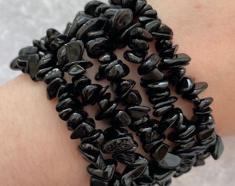 May include: A stack of black chip stone bracelets. The bracelets are made of small, irregularly shaped black stones strung together. The stones have a glossy finish and reflect light. The bracelets are worn on the wrist.
