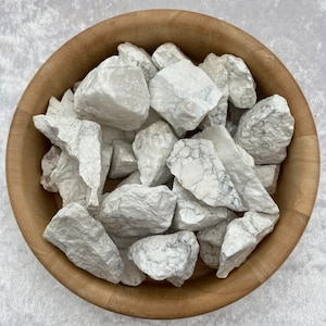 Könnte beinhalten: Eine Schale gefüllt mit weißen Howlith-Trommelsteinen. Die Steine sind rau und haben eine natürliche, weiße Farbe mit grauen Adern.