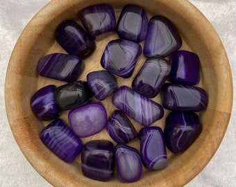 May include: A wooden bowl filled with purple agate tumbled stones. The stones have a variety of patterns and shades of purple.