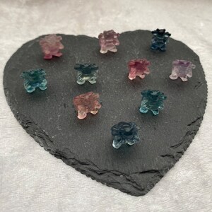 May include: A collection of ten small, carved crystal figurines in various shades of blue, pink, and purple. The figurines are arranged on a heart-shaped black slate surface.