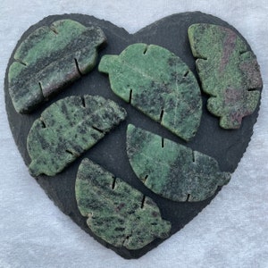 May include: Six green and black gemstone leaf-shaped carvings on a black slate heart-shaped surface.