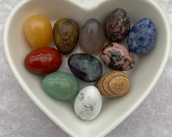 May include: A collection of ten smooth, polished, egg-shaped stones in various colors, including red, orange, yellow, green, blue, purple, brown, gray, and white, displayed in a white heart-shaped dish.