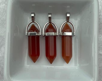 May include: Three carnelian crystal point pendants with silver-toned metal caps and loops. The pendants are displayed in a white ceramic dish.