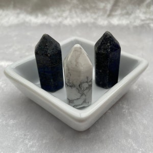 May include: Three crystal points, one white and two blue, are displayed on a white square dish. The points are arranged in a triangular pattern.