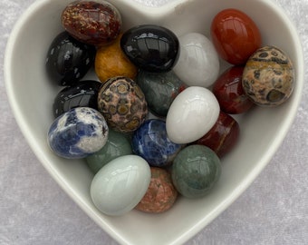 May include: A white heart-shaped bowl filled with various polished gemstone eggs. The eggs are in a variety of colors, including red, green, blue, black, white, and brown.