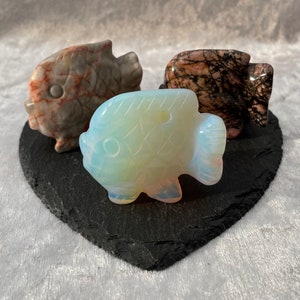 May include: Three carved fish figurines made of different types of stone. The fish on the left is a light brown with red and white streaks. The fish in the center is a light blue with white streaks. The fish on the right is a dark brown with pink and white streaks.