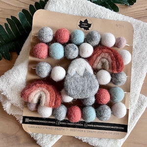 May include: A felt garland with a mountain, rainbow, and white, gray, and red balls. The garland is 7 feet long and is best for a 6 foot drape length or less. @bornandraisedcanada BORN + RAISED CANADA WWW.BORNANDRAISEDCAN.ETSY.COM