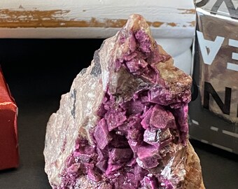 May include: A cluster of purple crystals embedded in a rough, gray rock. The crystals are sharp and have a metallic sheen.