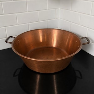 May include: A large, round copper pan with two small handles. The pan has a wide, shallow bowl and a shiny, reflective surface. The copper colour is a warm, reddish-brown. The pan is likely used for cooking or serving.