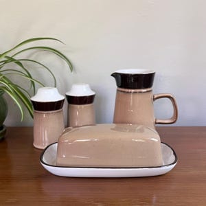 Könnte beinhalten: Ein Vintage-Keramik-Geschirrset in neutralen Farbtönen. Das Set umfasst eine Butterdose, einen Krug und zwei Salz- und Pfefferstreuer. Die Butterdose hat einen weißen Sockel mit schwarzem Rand. Krug und Streuer haben braune und weiße Akzente.