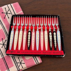 May include: A set of twelve two-pronged stainless steel cocktail forks with colorful plastic handles in light blue, white, pink, and black, featuring gold floral patterns. The box is labeled 18 STAINLESS-STEEL Special Quality.