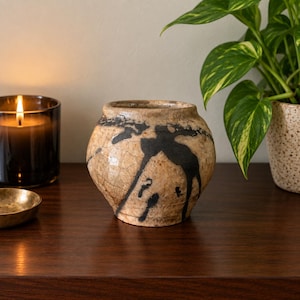 May include: A small, round ceramic pot with a beige, crackled glaze finish and bold, black abstract brushstroke patterns. The vessel features a wide, slightly tapered opening and a smooth, rounded body, showcasing a distinct, textured surface.