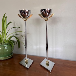May include: Two tall, silver-toned candle holders with a tulip-shaped design. The candle holders have a gold-colored accent at the base and the top. The base is a square shape. A green potted plant is in the background.