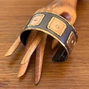 May include: A wide, open cuff bracelet with a dark grey band and gold trim. The bracelet features square copper-coloured inlays with gold borders and a central dot. The bracelet is displayed on a wooden hand.