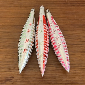 Pode incluir: Três enfeites de Natal alongados com glitter prateado e detalhes em vermelho. Cada enfeite tem um topo prateado e formato de gota. Os enfeites têm um padrão de formas de cruz vermelhas e linhas verticais. Os enfeites estão em uma superfície de madeira.