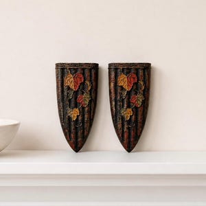 Puede incluir: Dos apliques de pared decorativos con forma de escudo. Los apliques son de color marrón oscuro con líneas verticales y presentan un diseño de vid con hojas rojas, amarillas y verdes. Se exhiben en una repisa blanca.