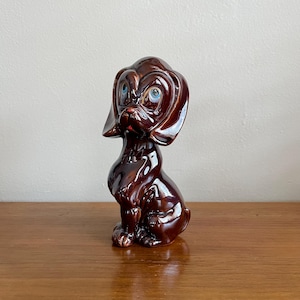 Könnte beinhalten: Eine braune Keramik-Hundefigur mit großen blauen Augen und einer roten Nase. Der Hund sitzt mit dem Kopf zur Seite geneigt.
