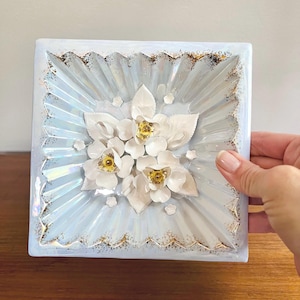 May include: Square, light blue decorative tile with a floral design. The tile features a cluster of white flowers with gold centers and gold trim around the edges. The background has a radial pattern.