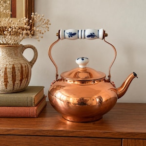 May include: A polished copper tea kettle with a rounded body and a curved spout. The kettle features a white ceramic handle and a matching lid knob, both decorated with blue floral patterns. The metal surface reflects the surrounding environment.