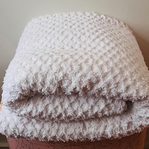 May include: A folded white blanket with a textured surface. The blanket has a raised, grid-like pattern and a soft, fluffy appearance. The blanket is folded over, revealing multiple layers.