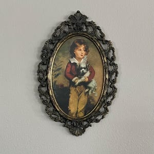 Puede incluir: Retrato ovalado enmarcado de un niño sosteniendo un perro pequeño. El retrato presenta a un niño con una chaqueta roja y pantalones color canela, con una camisa de cuello blanco. El marco es ornamentado, con un acabado bronce oscuro.
