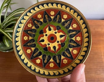 May include: A round, decorative plate with a detailed, colourful design. The plate features a central yellow circle with a red centre, surrounded by green, red, and black patterns. The outer edge is gold-coloured.