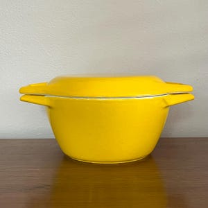 May include: A vibrant yellow casserole dish with a lid and side handles. The dish has a rounded form and a matching lid. The interior is white. The dish is on a wooden surface.