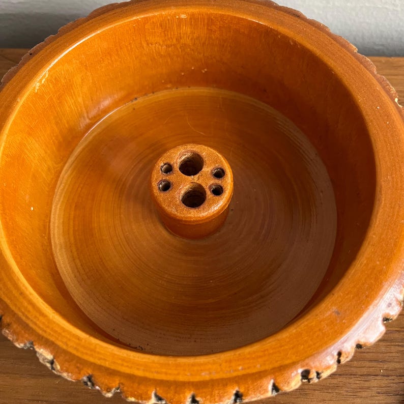 May include: A wooden bowl with a natural, light brown finish. The bowl has a central, cylindrical feature with multiple holes, and a textured rim. The wood grain is visible throughout the bowl's interior and exterior.