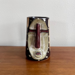 Könnte beinhalten: Eine Keramikvase mit einem Stammesmasken-Design. Die Vase hat einen dunkelbraunen Hintergrund mit einem beigefarbenen Gesicht und einer burgunderroten kreuzförmigen Nase. Weiße Punkte und beige vertikale Streifen verzieren die Seiten. Die Vase steht auf einer Holzoberfläche.