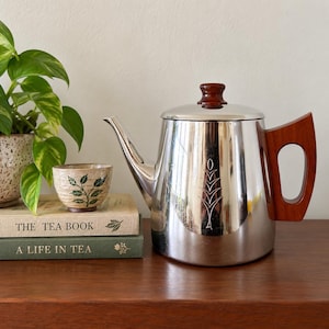 May include: A polished stainless steel teapot with a dark wood handle and lid knob, featuring a vertical etched leaf design. Beside it sits a speckled ceramic teacup with a green leaf pattern, resting on two books titled The Tea Book and A Life in Tea.