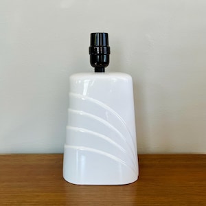 May include: A white ceramic lamp base with a black lamp holder. The base has a rounded rectangular shape with curved, raised lines on the front. The lamp sits on a wooden surface.