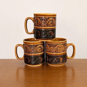 Può includere: Tre tazze in ceramica impilate, marroni e nere. Le tazze presentano un motivo strutturato di spirali e linee. Ogni tazza ha un manico. Sono esposte su una superficie di legno.