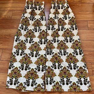 May include: Two fabric panels with a repeating floral design in brown, green, and burgundy on a cream backdrop. The fabric has a heavy weave, possibly for curtains or upholstery. The panels are displayed on a wooden surface.